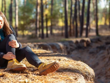 Frau sitzt im Wald auf dem Boden und hält sich den Fuss, schmerzverzerrtes Gesicht
