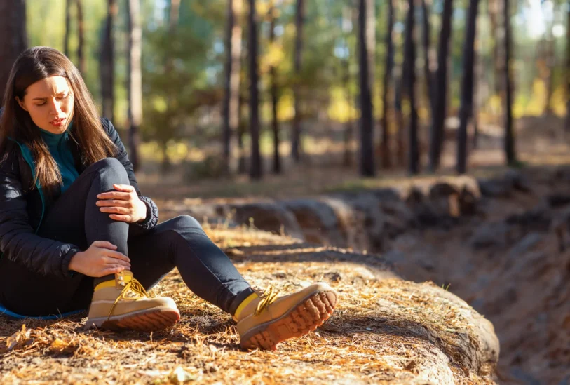 Frau sitzt im Wald auf dem Boden und hält sich den Fuss, schmerzverzerrtes Gesicht